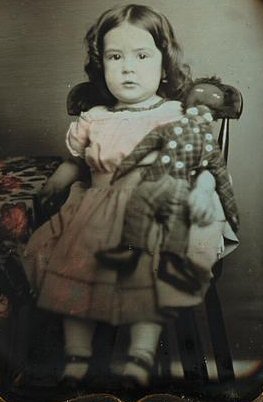Child seated beside table with table cloth holding a black rag doll
